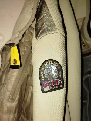 Beige vindjacka från Parajumpers - ÄKTA Snygg beige vindjacka från Parajumpers med patch på ärmen och coola detaljer i gult och orange. Jackan har quiltad front, hög krage och dragkedja. Materialet är en mix av slät och strukturerad textil för en sportig look. Perfekt för dig som gillar streetwear. Obs jackan är ganska sliten (litet brännhål, några färgfläckar på ena ärmen som går att få bort samt dragkedjan har gått av men finns kvar, går att laga även den.)