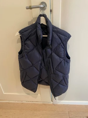 Mörkblå quiltad dunväst - Snygg mörkblå dunväst med hög krage och dragkedja framtill. Västen har ett quiltat mönster i stora rutor och är vadderad för extra värme. Perfekt att slänga över en hoodie eller tröja för en chill och trendig look.