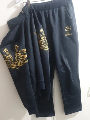 Mörkblå mjukisbyxor från Classilal - Mörkblå mjukisbyxor från Classilal med guldfärgat tryck och logga på vänster sida. Byxorna har resår i midjan och två sidofickor. Tillverkade i mjukt material som passar perfekt för chill eller träning. Snygg detalj med guldfärgade mönster.