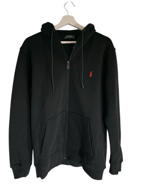 Svart Kofta från Polo Ralph Lauren - Klassisk svart hoodie med dragkedja från Polo Ralph Lauren. Tröjan har huva med dragsko, två fickor framtill och den ikoniska röda logotypen broderad på bröstet. Tillverkad i mjuk bomullsblandning och har ribbade muddar vid ärmslut och nederkant.