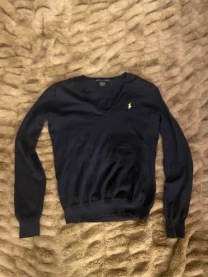 Mörkblå stickad tröja från Ralph Lauren - Mörkblå stickad tröja från Ralph Lauren med v-ringning och klassisk gul broderad logga på bröstet. Tröjan har långa ärmar och ribbade muddar vid ärmslut och nederkant. Perfekt för en stilren och tidlös look.