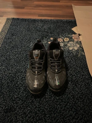 Nike Air Vapormax svarta sneakers - Säljer ett par svarta Nike Air Vapormax sneakers med reflekterande detaljer och en grön swoosh på sidan. Skorna har en låg silhuett och genomskinlig sula med synliga luftbubblor. Perfekta för dig som gillar sportig och modern stil.