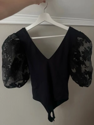 Svart body med puffärmar från Zara - Trendig svart body från Zara med djup V-ringning och stora puffärmar i transparent mesh med blommigt mönster. Kroppsnära passform och knäppning nedtill. Perfekt för dig som vill ha en statement-piece i garderoben.