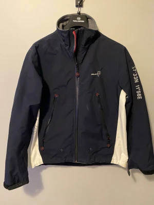 Marinblå vindjacka från Pelle P - Snygg marinblå vindjacka från Pelle P med vita paneler på sidorna och diskreta röda detaljer. Jackan har hög krage, två dragkedjeförsedda fickor framtill och tryckt koordinattext på ena ärmen. Materialet är slitstarkt och vattenavvisande, perfekt för blåsiga dagar. Riktigt fräsch och snygg för de som vill komponera grisch stilen med en vindjacka, denna jacka är väldigt varm innuti så du kan använda den när de kallt ute också 