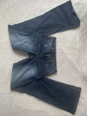Mörkblå bootcut jeans med slitningar - Säljer ett par mörkblå bootcut jeans med snygga slitningar och tvättade detaljer framtill. Klassisk femficksmodell med dragkedja och knapp. Jeansen har en avslappnad passform och är tillverkade i denim.