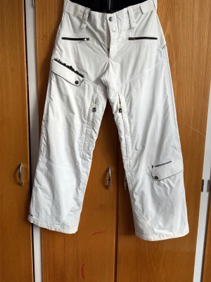 Peak performance Vita täckbyxor  - Säljer ett par vita täckbyxor från Peak Performance med raka ben, flera dragkedjeförsedda fickor och knappstängning i midjan. Byxorna har förstärkta partier och är perfekta för vinterns alla äventyr. Materialet är slitstarkt och vattenavvisande.
