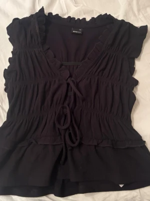 Svart blus med snörning från Stradivarius - Supersöt svart blus från Stradivarius med volangkantad v-ringning och snörning framtill. Blusen har korta ärmar och en lätt rynkad struktur som ger en feminin siluett. Perfekt att styla med jeans eller kjol för en trendig look.