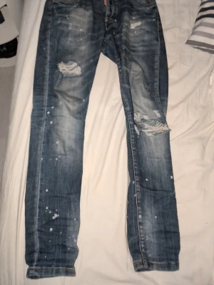Blå slitna jeans från Dsquared2 - Säljer ett par blå jeans från Dsquared2 med slitningar, hål och färgstänk för en riktigt trendig och avslappnad look. Jeansen har raka ben och klassisk femficksmodell. Perfekta för dig som gillar streetstyle och vill sticka ut.