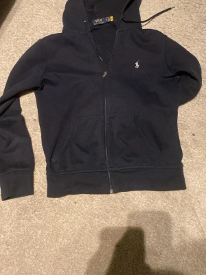 Marinblå hoodie från Polo Ralph Lauren - Snygg marinblå hoodie från Polo Ralph Lauren med dragkedja framtill och klassisk logga broderad på bröstet. Hoodien har huva med dragsko och två fickor framtill. Tillverkad i mjukt bomullsmaterial som är skönt att ha på sig. 