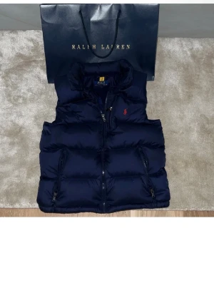 Mörkblå dunväst från Polo Ralph Lauren - Snygg mörkblå dunväst från Polo Ralph Lauren med hög krage, dragkedja framtill och två fickor med dragkedja. Klassisk röd logga på bröstet och quiltad design som ger en puffig look. Perfekt för lager på lager och streetstyle.
