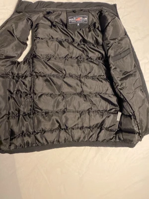 Svart dunväst från Polo Club - Snygg svart dunväst från Polo Club Saint Vincent. Västen har quiltad design och är tillverkad i glansigt material som ger en modern look. Perfekt att slänga över en hoodie eller tröja för extra värme. Märkeslapp på insidan och klassisk passform.