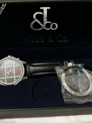 Jacob & Co. silverfärgad klocka - Snygg klocka från Jacob & Co. med silverfärgat ur och glittrande stenar på urtavlan. Svart läderarmband och klassisk rund form. Kommer i originalask och med extra ram. Perfekt för dig som gillar exklusiva accessoarer.