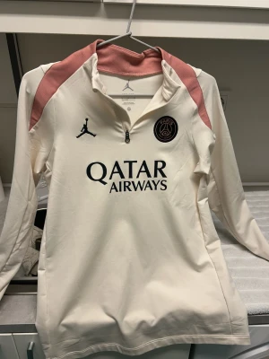 PSG träningströja Jordan x Qatar - Snygg långärmad träningströja från PSG i samarbete med Jordan. Tröjan är cremevit med rosa detaljer på axlarna, har halv dragkedja och PSG-logga samt Jordan-logga på bröstet. Qatar Airways tryckt på framsidan. Perfekt för fotbollsträning eller chill.