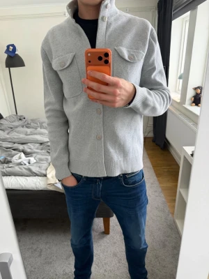 Ljusgrå cardigan fån Less Worries - Snygg ljusgrå cardigan med knäppning framtill och två bröstfickor med lock och knapp. Tröjan har ribbade muddar vid ärmslut och nederkant samt hög krage. Perfekt att slänga på sig över en t-shirt för en clean look.