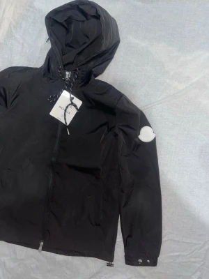 Svart vindjacka från Moncler - Snygg svart vindjacka från Moncler med huva och dragkedja framtill. Jackan har Moncler-logga på ärmen och justerbara muddar med tryckknappar. Perfekt för dig som vill ha en stilren och funktionell jacka.