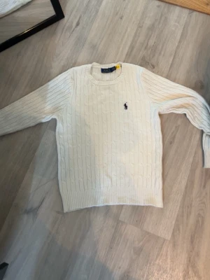 Vit stickad tröja Polo Ralph Lauren - stickad vit tröja från Polo Ralph Lauren med rund halsringning och den klassiska broderade loggan på bröstet. Tröjan har långa ärmar och ribbade muddar vid ärmslut och nederkant. Perfekt för en clean och stilren look.
