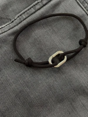 Svart armband - Stilrent svart armband i justerbar textilsnodd med en silverfärgad metallhake som detalj. Perfekt för dig som gillar minimalistisk och modern stil. Passar både ensam eller i kombination med andra armband.
