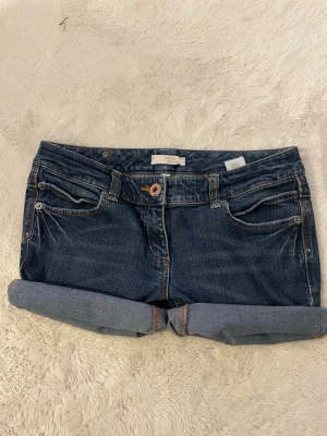 Mörkblå jeansshorts med uppvik - Säljer ett par mörkblå jeansshorts från True Religion med uppvikta benslut och snygga kontrastsömmar. Shortsen har fem fickor, bälteshällor och klassisk knappgylf. Bakfickorna har dekorativa sömmar och en knappdetalj.