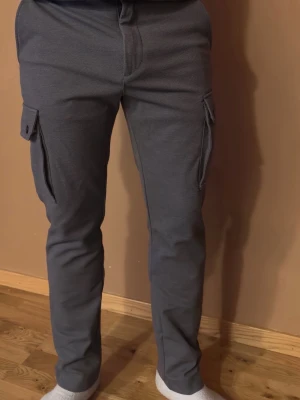  cargobyxor passar S/M - Säljer ett par mörkgrå cargobyxor med raka ben och stora sidofickor med lock och knapp. Byxorna har en modern, avslappnad passform och är tillverkade i ett mjukt, stretchigt material som är skönt att bära. Perfekta för en casual streetwear-look.