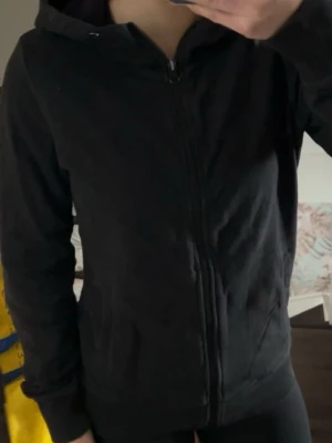 Svart zip hoodie med huva - En enkel och stilren svart zip hoodie med dragkedja framtill och två fickor. Hoodien har en klassisk huva och ribbade muddar vid ärmsluten. Perfekt för en avslappnad look och funkar året runt. Materialet känns mjukt och bekvämt.