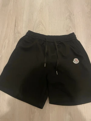 Svarta shorts från Moncler med snörning - Säljer ett par svarta shorts från Moncler med elastisk midja och snörning framtill. Shortsen har Moncler-loggan på vänster ben och är tillverkade i mjuk bomull, perfekta för en avslappnad stil.