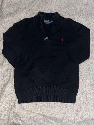 Svart halvzip från Polo Ralph Lauren - Klassisk svart tröja med halv dragkedja från Polo Ralph Lauren. Tröjan har ribbade muddar, hög krage och den ikoniska röda logotypen broderad på bröstet. Perfekt för en stilren och avslappnad look.