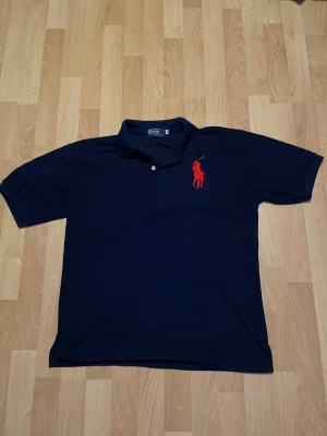 Mörkblå pikétopp från Polo Ralph Lauren - Mörkblå pikétopp från Polo Ralph Lauren med klassisk krage och två knappar vid halsen. Stor röd broderad logga på bröstet. Tillverkad i 100% bomull och har korta ärmar för en clean och sportig look.