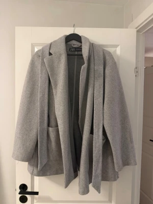 Ljusgrå kappa från Zara med skärp - Stilren ljusgrå kappa från Zara med klassisk krage och knytskärp i midjan. Kappan har två stora fickor framtill och en rak, avslappnad passform. 