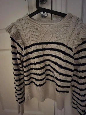 Randig stickad tröja med volang - Säljer en beige stickad tröja med mörkblå ränder och volangdetaljer vid axlarna. Tröjan har kabelstickade partier och ett snyggt mönster framtill. Långa ärmar och ribbade muddar ger en skön passform. Perfekt för dig som gillar en trendig och unik look.