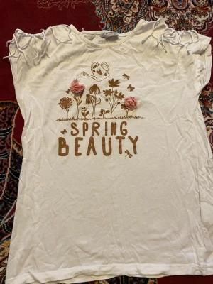 Vit t-shirt med blommor  - Vit t-shirt från LC Waikiki med tryck av blommor och texten 'Spring Beauty' framtill. T-shirten har korta ärmar och dekorativa fransar vid axlarna. Materialet känns mjukt och luftigt, perfekt för vår och sommar.