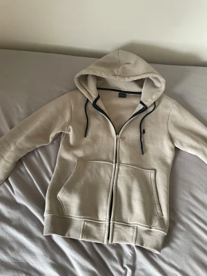 Beige hoodie från Polo Ralph Lauren - Snygg beige hoodie från Polo Ralph Lauren med dragkedja framtill och två stora fickor. Har svart logga broderad på bröstet och svarta snören i huvan. Tillverkad i mjukt bomullsmaterial och har ribbade muddar vid ärmar och nederkant. (Funkar som s och m)