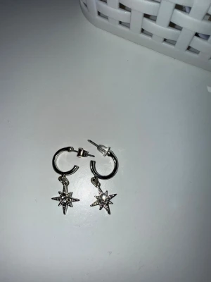 Silvriga örhängen med stjärnhänge - Säljer ett par silvriga örhängen i ringmodell med hängande stjärnor dekorerade med små glittrande stenar. Örhängena har en modern och trendig design som passar perfekt till dig som gillar detaljer. Materialet ser ut att vara metall med stenar som ger extra bling.