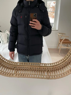 Svart pufferjacka från Polo Ralph Lauren - Säljer en marinblå pufferjacka från Polo Ralph Lauren med huva och dragkedja. Jackan har det klassiska röda Polo-logot broderat på bröstet och flera praktiska fickor med dragkedja. Perfekt för kalla dagar och riktigt snygg till Stockholm-stilen eller grish stilen