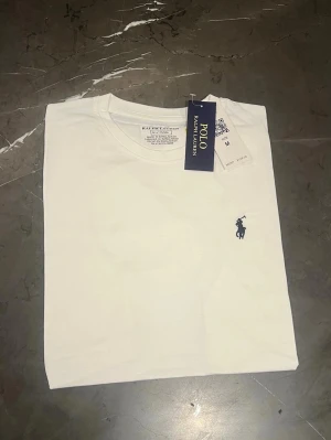 Vit t-shirt från Polo Ralph Lauren - Klassisk vit t-shirt från Polo Ralph Lauren med rund hals och korta ärmar. Diskret mörkblå logga broderad på bröstet. Tillverkad i mjuk bomull för en skön och clean look. Perfekt basplagg till garderoben.