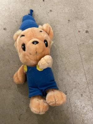Bamse mjukisdjur i blå kläder - Säljer ett gulligt Bamse mjukisdjur med blå overall och blå luva. Mjukdjuret är beige med svarta broderade ögon och näsa samt gula detaljer på axlarna. Perfekt för dig som gillar klassiska seriefigurer!