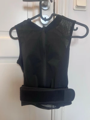 Svart skyddsväst för sport, stl 134-152 - Svart skyddsväst med dragkedja framtill och bred kardborreknäppning i midjan som går att korrigera upp och ner. Västen är tillverkad i ett ventilerande meshmaterial och passar perfekt för sporter där extra skydd behövs, som skidåkning eller cykling. Snygg och funktionell design utan ärmar. Totalt använt 2veckor.