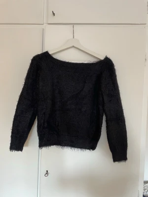 Svart fluffig bandeau tröja - Mysig svart stickad bandeau tröja med fluffig textur. Tröjan har långa ärmar och en avslappnad passform, perfekt för att hålla värmen under kyliga dagar. Passar till både jeans och kjol för en trendig look. Extremt mjuk och skön