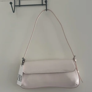 Ljusrosa axelväska från Zara - Supersnygg ljusrosa axelväska från Zara med stilren design och smal axelrem. Väskan är tillverkad i syntetmaterial med lätt glansig finish och har ett lock som stängs framtill. Perfekt för att lyfta din outfit med en fräsch färg.