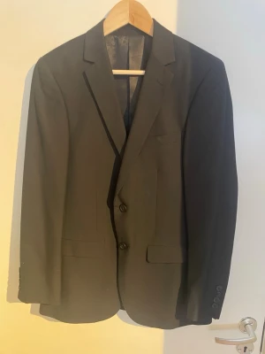 Svart slim fit kavaj Dressmann - Snygg svart kavaj från Dressmann i slim fit-modell. Klassisk design med två knappar framtill, tre fickor och slät finish. Perfekt för dig som vill ha en stilren och modern look. 