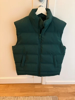 Mörkgrön dunväst med hög krage från Weekday - Stilren mörkgrön dunväst med hög krage och dragkedja framtill. Västen har två snedställda fickor och breda quiltade paneler för extra värme. Perfekt att slänga över en hoodie eller tröja för en cool lager-på-lager-look.