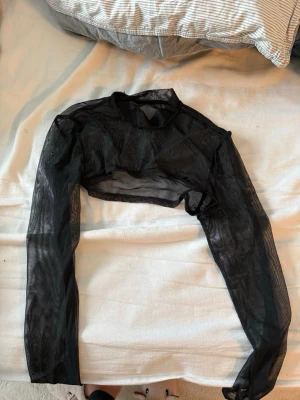 Svart mesh bolerotopp - Säljer en svart mesh bolerotopp med lång ärm och croppad passform. Toppen är genomskinlig och har en stilren, edgy look som passar perfekt över ett linne eller klänning. Perfekt för att addera lager till din outfit.