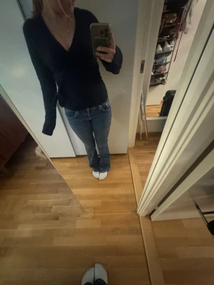Gina tricot tröja - Säljer en mörkblå stickad omlottopp med djup v-ringning och långa ärmar. Tröjan har en figurnära passform och knyts i sidan för en snygg siluett. Perfekt att matcha med jeans eller kjol för en trendig look.