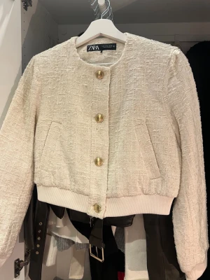 Beige bouclékofta från Zara - Säljer en beige bouclékofta från Zara med rund halsringning och stora guldfärgade knappar framtill. Koftan har ribbade muddar vid ärmslut och nederkant samt två fickor fram. Perfekt för dig som gillar en chic och trendig look.