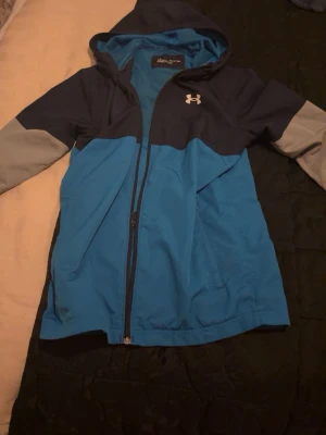 Blå vindjacka från Under Armour - Snygg vindjacka från Under Armour i blått med mörkblå axelparti och grå ärmar. Jackan har huva, dragkedja framtill och två sidofickor. Perfekt för blåsiga dagar och sportiga outfits. Tillverkad i lätt och tåligt material.
