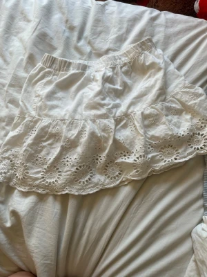 Vit volangkjol med broderade detaljer - Söt vit kjol med volang och broderade blommor längs nederkanten. Kjolen har resår i midjan och är gjord i ett lätt bomullstyg som känns luftigt och somrigt. Perfekt för dig som gillar romantisk och feminin stil.