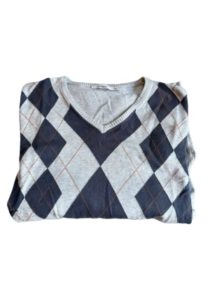 Grå argyle stickad tröja golf - Snygg stickad tröja med klassiskt argylemönster i grått, svart och tunna röda linjer. V-ringad halsringning och långa ärmar. Perfekt för dig som gillar retrovibbar och vill ha en unik look. Materialet känns mjukt och bekvämt.