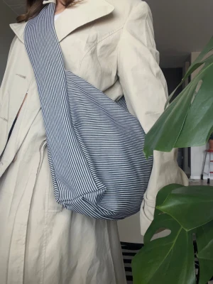 Randig svart axelväska i tyg - Säljer en stor axelväska i blått tyg med tunna vita ränder. Väskan har en bred axelrem och rymlig form, perfekt för att bära med sig allt man behöver. Enkel och stilren design som passar till många outfits.