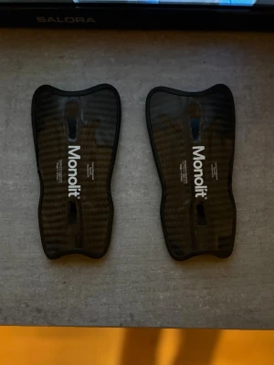 Monolit benskydd i kolfiber - Säljer ett par svarta benskydd från Monolit i kolfiber. De har en slimmad och ergonomisk form med vit logga på framsidan. Insidan är vadderad för extra komfort och skydd. Perfekta för dig som vill ha lätta och tåliga benskydd till träning eller match.