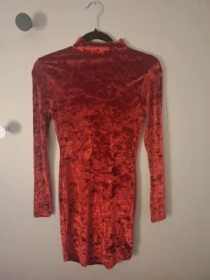 Röd sammetsklänning Gina Tricot - Snygg röd sammetsklänning från Gina Tricot i kort modell. Klänningen har lång ärm och hög krage, vilket ger en trendig och elegant look. Materialet är mjukt och glansigt sammet som känns lyxigt och bekvämt.