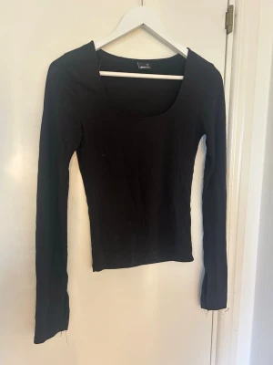 Svart långärmad topp från Gina Tricot - En enkel och stilren svart långärmad topp från Gina Tricot med rundad, djup urringning. Modellen är tight och passar perfekt till många olika looks. Tillverkad i mjukt bomullsmaterial som känns skönt mot huden.